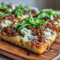 Une pizza flatbread croustillante garnie d'oignons caramélisés sucrés, de fromage crémeux, d'arugula fraîche et d'un filet de réduction balsamique acidulée—idéale en apéritif gourmet ou plat principal léger.